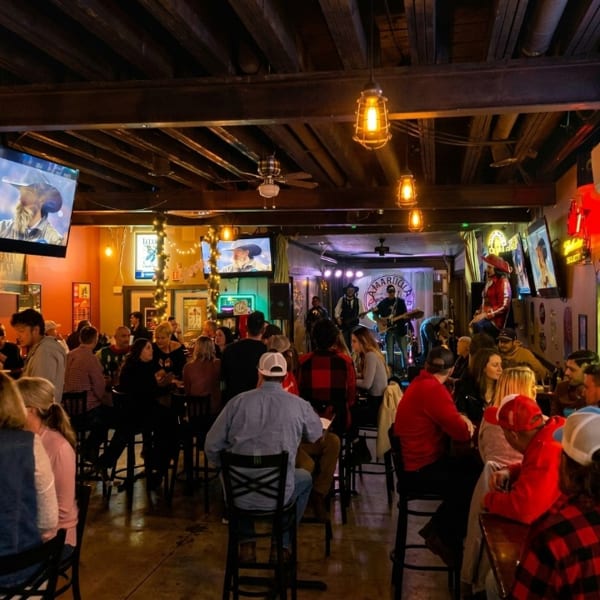 A lively bar scene with a band performing on stage and patrons seated at high-top tables.