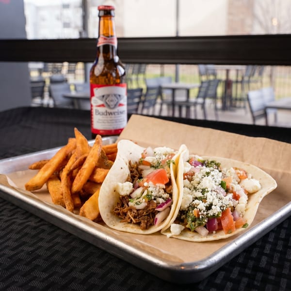 Two tacos with fries and a Budweiser beer on a metal tray, with an outdoor patio in the background.