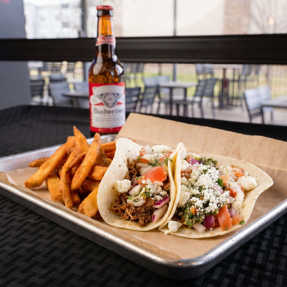 Two tacos with fries and a Budweiser beer on a metal tray, with an outdoor patio in the background.