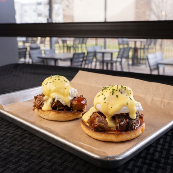Two eggs Benedict with pulled pork and hollandaise sauce on a tray, with an outdoor patio in the background.