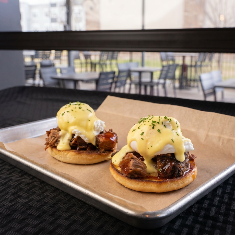 Two eggs Benedict with pulled pork and hollandaise sauce on a tray, with an outdoor patio in the background.