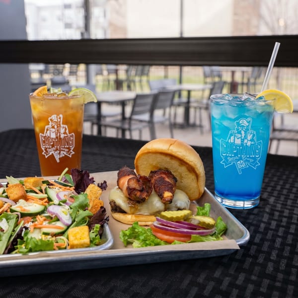 A burger with bacon-wrapped jalapeños, a salad, and two Captains Sports Lounge drinks on a tray.